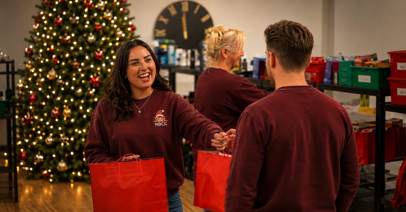 NBCC volunteers in maroon branded sweatshirts sharing a laugh as they hand over Red Bags Full of Joy by the Christmas tree at the Elves Workshop